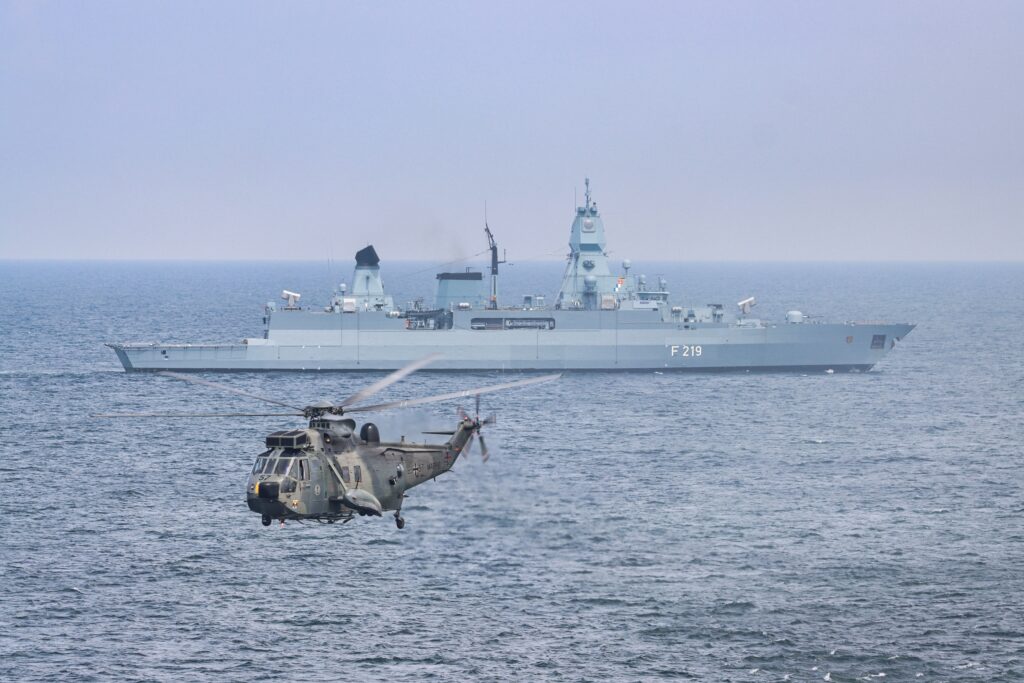 A military helicopter flies over the sea near a German warship A military helicopter flies over the sea near a German warship